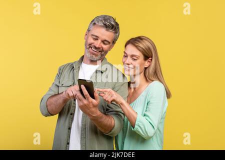 Excited caucasian middle aged couple using cellphone and pointing on it, looking at smartphone screen and smiling Stock Photo