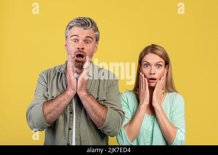 Omg. Shocked middle aged spouses touching face and cheeks, looking at camera isolated on yellow studio background Stock Photo