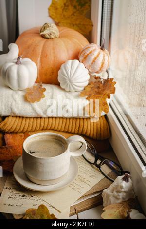 Cozy autumn morning still life scene. Steaming cup of hot coffee near the window. Fall, Thanksgiving concept. Pumpkins and leaves, wool sweater Stock Photo