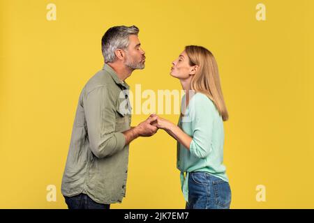 Middle aged couple in love pursing lips for kiss over yellow studio background, profile side view Stock Photo