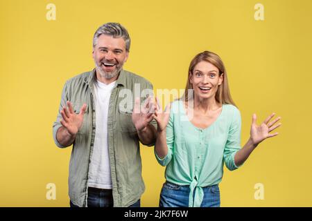 Amazed middle aged spouses expressing amazement, screaming and showing their palms, gesturing over yellow background Stock Photo