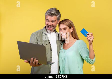 Overjoyed mature man and woman using credit card and laptop, standing over yellow background, studio shot Stock Photo