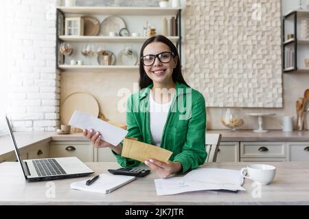 Happy and successful business woman working at home in kitchen, looking at camera and smiling wearing green magpie, brunette holding letter and document Stock Photo