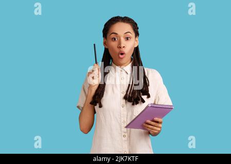 Portrait of inspired joyful woman with dreadlocks looking amazed with sudden genius idea and holding pencil to write in notebook, wearing white shirt. Indoor studio shot isolated on blue background. Stock Photo