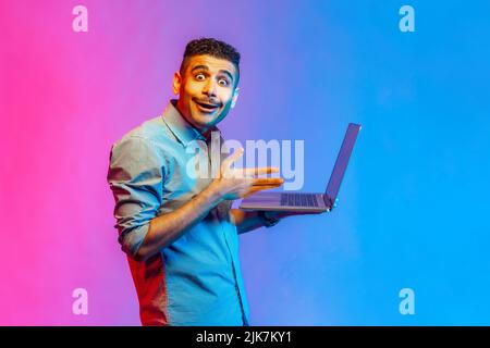 Portrait of amazed handsome man in shirt watching shocked video, having astonished facial expression, looking at camera with big eyes. Indoor studio shot isolated on colorful neon light background. Stock Photo