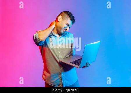 Portrait of tired sick man freelancer in shirt holding his neck after hard overtime work and make massage of neck pain. Indoor studio shot isolated on colorful neon light background. Stock Photo