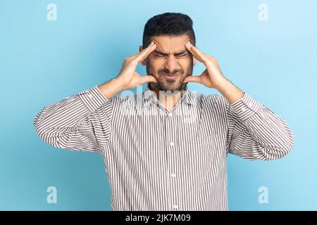 Unhappy frustrated bearded businessman massaging temples feeling headache, suffering migraine or high blood pressure, wearing striped shirt. Indoor studio shot isolated on blue background. Stock Photo
