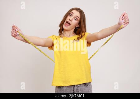 Beautiful overjoyed teen girl in yellow T-shirt holding tape line on her waist, measuring centimeters of body, keeping healthy lifestyle, workout. Indoor studio shot isolated on gray background. Stock Photo