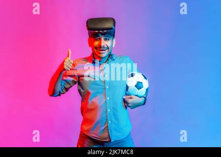 Portrait of man in shirt wearing virtual reality glasses, standing with soccer ball, showing thumb up, like football match. Indoor studio shot isolated on colorful neon light background. Stock Photo