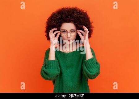 Portrait of attentive beautiful woman with Afro hairstyle wearing green casual style sweater standing holding glasses and looking at camera. Indoor studio shot isolated on orange background. Stock Photo