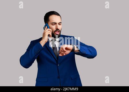 Portrait of handsome bearded man talking phone and looking at wristwatch, making appointment, waiting for date, wearing official style suit. Indoor studio shot isolated on gray background. Stock Photo