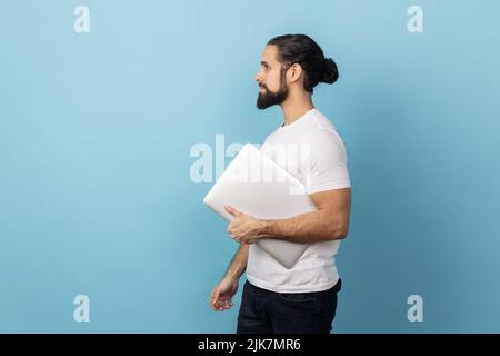 Side view portrait of man with beard wearing white T-shirt standing, holding closed laptop or folder, looking ahead, concentrated expression. Indoor studio shot isolated on blue background. Stock Photo