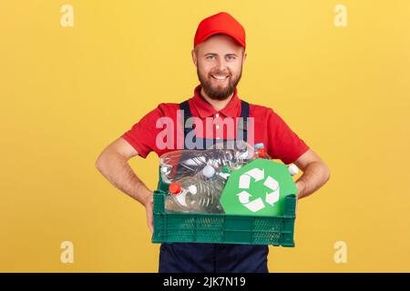 Portrait of happy bearded worker man standing and holding box with plastic bottles and recycling sign, wearing overalls and red cap. Indoor studio shot isolated on yellow background. Stock Photo