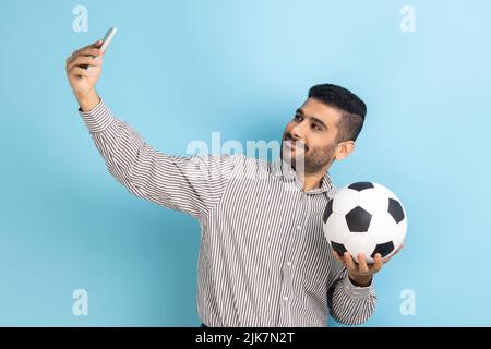 Young adult attractive businessman making selfie, having video call or broadcasting livestream with ball in hand, wearing striped shirt. Indoor studio shot isolated on blue background. Stock Photo