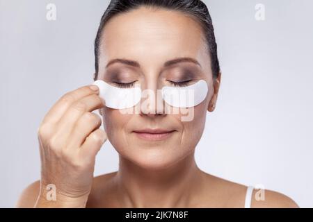 Closeup portrait of middle aged woman with cosmetic collagen patches under eyes, enjoys flawless of skin, has well cared complexion. Indoor studio shot isolated on gray background. Stock Photo