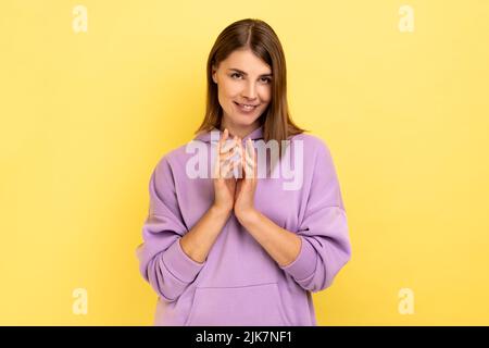 Portrait of devious cunning young woman clasping hands and smirking mysteriously, scheming cheats, evil prank, wearing purple hoodie. Indoor studio shot isolated on yellow background. Stock Photo