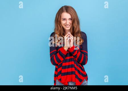 Portrait of young devious woman wearing striped casual style sweater looking at camera with cunning tricky face and smirk, planning evil trick. Indoor studio shot isolated on blue background. Stock Photo
