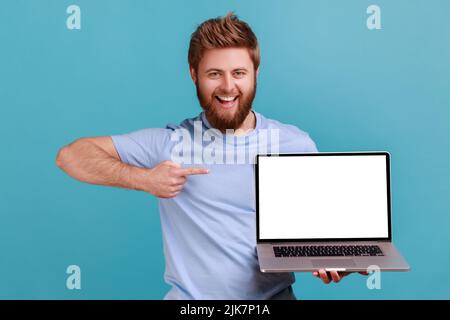 Portrait of happy bearded man holding laptop and pointing at empty screen, smiling satisfied with computer app, looking at camera. Indoor studio shot isolated on blue background. Stock Photo
