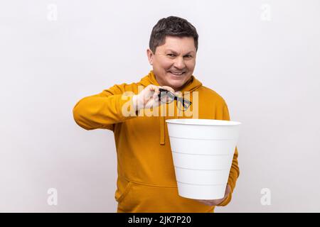 Portrait of happy satisfied middle aged man throwing out his optical glasses after vision treatment, looking at camera, wearing urban style hoodie. Indoor studio shot isolated on white background. Stock Photo