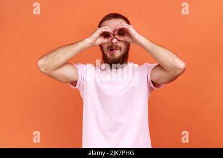Portrait of bearded man with funny face looking through fingers shaped like binoculars and expressing positive, wearing pink T-shirt. Indoor studio shot isolated on orange background. Stock Photo