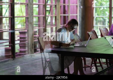 Young focused woman student wearing glasses making notes, studying remotely in cafe Stock Photo