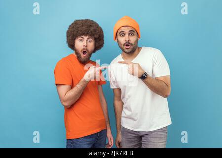 Portrait of two young adult hipster men pointing finger each other and looking at camera with shocked facial expressions and open mouth. Indoor studio shot isolated on blue background. Stock Photo