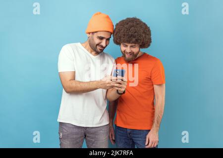 Portrait of two handsome optimistic young adult hipster men standing, using smartphone, reading positive news in social networks, smiling. Indoor studio shot isolated on blue background. Stock Photo