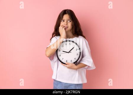 Portrait of nervous anxious little girl wearing white T-shirt biting nails on fingers holding big wall clock in hand, worried about deadline. Indoor studio shot isolated on pink background. Stock Photo