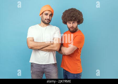 Portrait of two handsome serious young adult hipster men standing looking at camera with assertive expression, keeping hands folded. Indoor studio shot isolated on blue background. Stock Photo