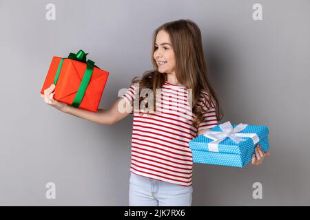 Portrait of charming little girl wearing striped T-shirt celebrating her birthday, holding two present boxes in hands, being happy with gifts. Indoor studio shot isolated on gray background. Stock Photo