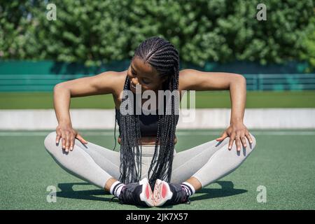African American woman sits on green track at sports ground stretching legs on sunny summer day Stock Photo