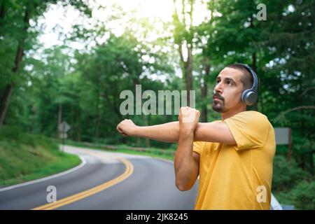 Young fitness man runner with earphones stretching before run in road in nature. Healthy lifestyle and sport concepts Stock Photo
