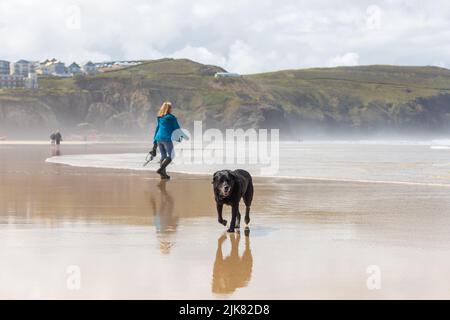 A black dog walks in wet sand toward the viewer on a beach as a lady passes by behind. Cornish cliffs, houses and sea background Stock Photo