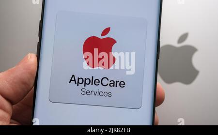 Apple Care Services logo seen on smartphone hold in a hand above the top of macbook with blurred apple logo. Concept. Stafford, United Kingdom, July 3 Stock Photo