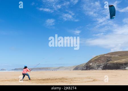 A lady flies a power kite on the beach with the kite soaring through the blue summer sky. Cliffs and sea behind. Stock Photo