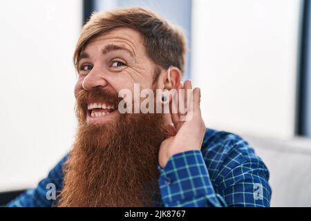 Young redhead man using deafness aid machine sitting on sofa at home Stock Photo