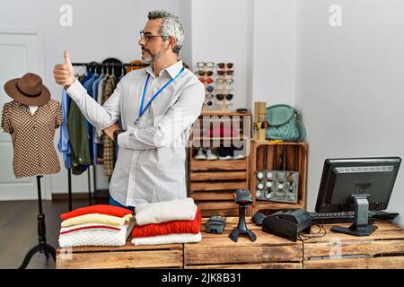 Middle age hispanic man working as manager at retail boutique looking proud, smiling doing thumbs up gesture to the side Stock Photo