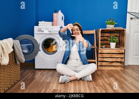 Hispanic woman doing laundry sitting on the floor smiling making frame with hands and fingers with happy face. creativity and photography concept. Stock Photo