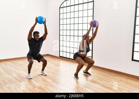 Young african american sporty couple doing squat exercise using ball at sport center. Stock Photo