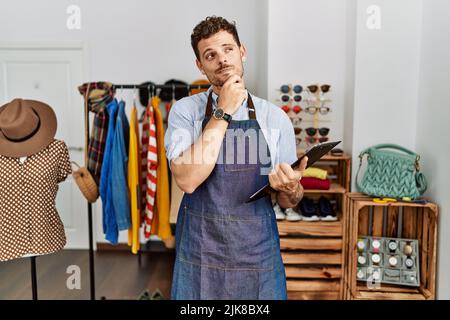 Handsome young man working as manager at retail boutique with hand on chin thinking about question, pensive expression. smiling with thoughtful face. Stock Photo