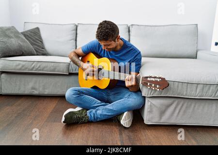 Young hispanic man playing guitar sitting on the floor at home Stock Photo