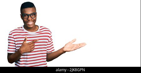 Young african american man wearing casual t shirt and glasses amazed and smiling to the camera while presenting with hand and pointing with finger. Stock Photo