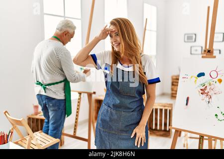 Hispanic woman wearing apron at art studio very happy and smiling looking far away with hand over head. searching concept. Stock Photo