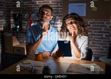Middle age hispanic couple using touchpad sitting on the table at night thinking concentrated about doubt with finger on chin and looking up wondering Stock Photo