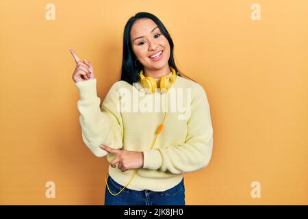 Beautiful hispanic woman with nose piercing wearing headphones on neck smiling happy pointing with hand and finger to the side Stock Photo