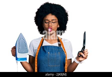 Young african american woman dressmaker designer wearing atelier apron holding iron and scissors making fish face with mouth and squinting eyes, crazy Stock Photo