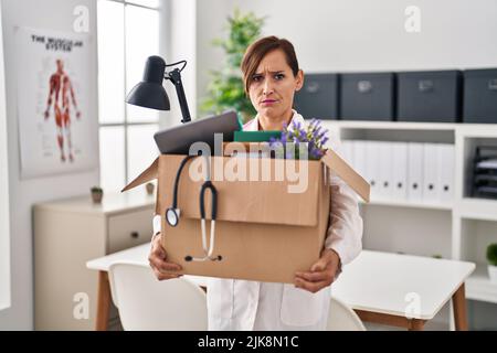 Middle age doctor woman holding cardboard box with items at the clinic skeptic and nervous, frowning upset because of problem. negative person. Stock Photo