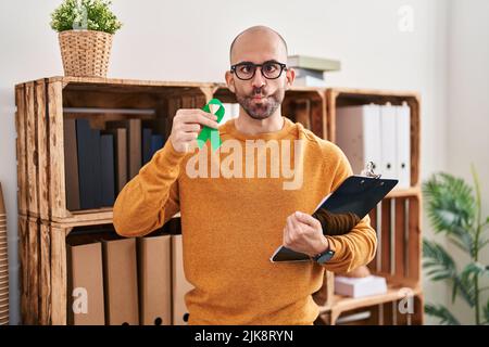 Young bald man with beard holding support green ribbon making fish face with mouth and squinting eyes, crazy and comical. Stock Photo