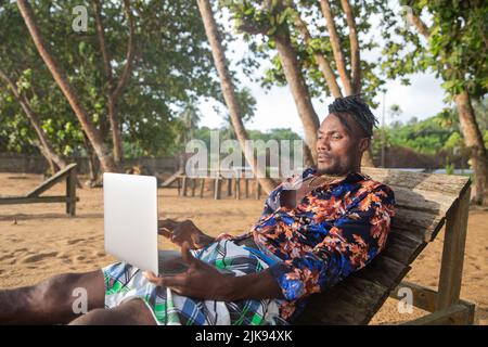 A digital nomad consults his computer while lying on the bench by the sea. Stock Photo