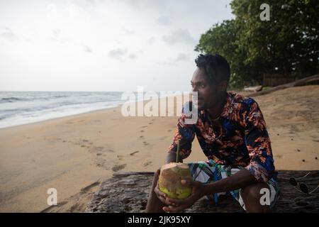 A black man sitting on a tree trunk by the sea enjoys his vacation by enjoying coco juice. Stock Photo
